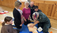 A group of people participates in a CPR training session, observing as an instructor demonstrates on a mannequin.