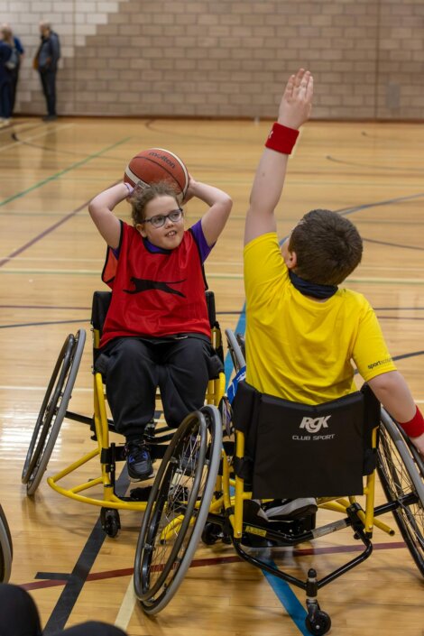 Two children in wheelchairs play basketball indoors. One child in a red jersey holds the ball overhead, while the other in a yellow jersey raises an arm to block.