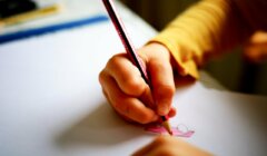 A child’s hand drawing on paper with a pencil, coloring a pink shape, wearing a yellow long-sleeve shirt.