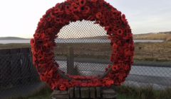 A large wreath made of red poppies is displayed on a stone pedestal in front of a chain-link fence, with a rural landscape in the background.