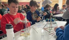 Students sit at tables in a classroom, constructing small structures with straws and clay.