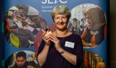 A person holding an award stands in front of a Scottish Library and Information Council backdrop, featuring diverse people engaged in library activities.