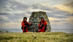 Two people in orange gear kneel by a stone monument on a grassy area near the sea, under a cloudy sky.