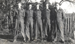 Five men in military uniforms stand in a row outdoors, in front of a fence and trees, posing for a group photo.