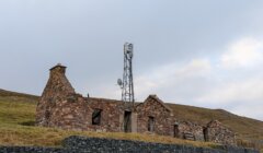 Abandoned stone building with a tall metal tower, set on a grassy hillside under a cloudy sky.