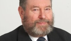 An older man with a gray beard wearing a suit, tie, and lapel pin smiles slightly against a plain background.