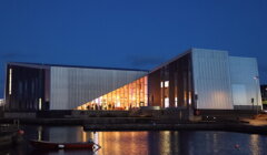 Modern waterfront building with a geometric facade illuminated from within at dusk, reflecting on the water. A small boat is moored in the foreground.