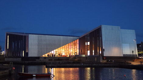 Modern waterfront building with a geometric facade illuminated from within at dusk, reflecting on the water. A small boat is moored in the foreground.