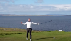 A person stands on a grassy golf course holding a club, with a lake and distant hills in the background under a partly cloudy sky.