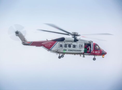 A red and white HM Coastguard helicopter in flight with visible spinning rotors and the side door open.