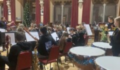 An orchestra rehearses in a hall with red curtains and a decorated Christmas tree. Musicians are seated with instruments and sheet music stands.