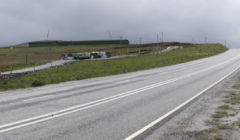 A wet, empty road curves through a grassy landscape with scattered construction materials and a wind turbine in the distance under a cloudy sky.