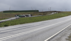 A wet, empty road curves through a grassy landscape with scattered construction materials and a wind turbine in the distance under a cloudy sky.