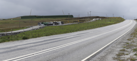 A wet, empty road curves through a grassy landscape with scattered construction materials and a wind turbine in the distance under a cloudy sky.