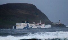 Ferry with "NorthLink" logo navigates rough seas near a rocky coastline and lighthouse.