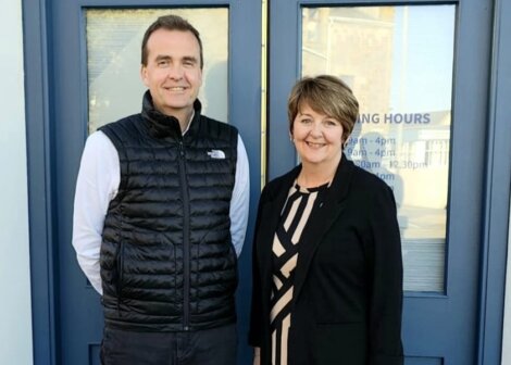 Two people stand smiling in front of a blue door with opening hours listed on it. The man is wearing a black vest and white shirt, and the woman is in a black outfit with a patterned blouse.