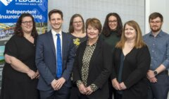 A group of seven people dressed in business attire standing in front of a banner that reads "TL Dallas Independent Insurance Broking.