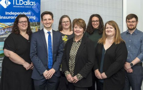 A group of seven people dressed in business attire standing in front of a banner that reads "TL Dallas Independent Insurance Broking.