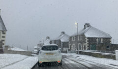 A car drives on a snow-covered road lined with stone houses against a gray sky. Snow lightly falls, and streetlights illuminate the scene.