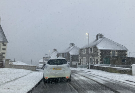 A car drives on a snow-covered road lined with stone houses against a gray sky. Snow lightly falls, and streetlights illuminate the scene.