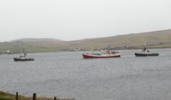 A red and white boat is escorted by two smaller tugboats in a grey, misty harbor, with hills and a few buildings in the background.