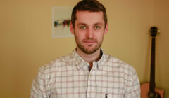 A man wearing a plaid shirt stands in a room with light-colored walls. A guitar leans against the wall in the background.