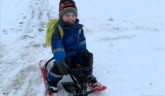 A child in a blue winter outfit and colorful hat is sitting on a red sled in the snow, smiling.