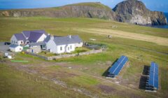 Aerial view of a rural area with houses, solar panels, grassland, and a large rock formation by the sea.