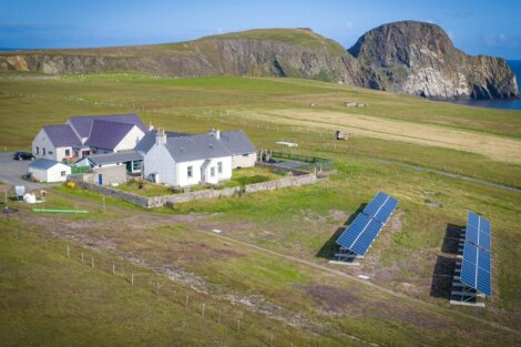 Aerial view of a rural area with houses, solar panels, grassland, and a large rock formation by the sea.