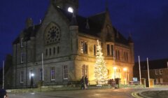 A historic stone building at night with a large, illuminated Christmas tree in front, framed by glowing windows and streetlights.