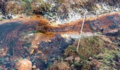 A narrow, polluted stream runs through grass and soil, showing orange and black discoloration, with a thin wooden stick placed upright nearby.