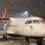 A white propeller plane is parked on a snowy tarmac at night, with snow falling and airport lights in the background.