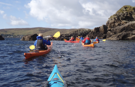 People kayaking in a line on open water near rocky cliffs, with a cloudy sky and hilly landscape in the background.