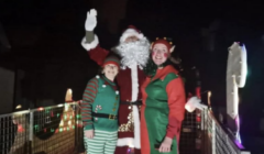 Santa and two elves wave from a festively lit vehicle at night.