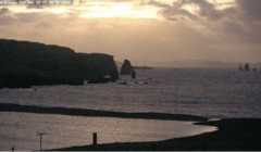 A rocky coastline at sunrise with scattered clouds in the sky, casting reflections on the water and silhouetting the cliffs and sea stacks.