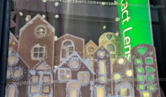 Gingerbread-style houses with white icing details are displayed in a shop window. Snowflakes and a green sign with "contact lenses" are visible.