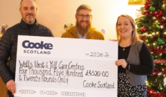 Three people hold a large check for £4,520.00 from Cooke Scotland to Walls, Unst & Yell Care Centres. A Christmas tree is in the background.