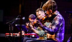 Three musicians performing on stage with violins and a keyboard.