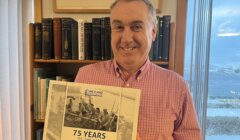 Man holding a "75 Years of the SFA" book, standing in front of a bookshelf and window.