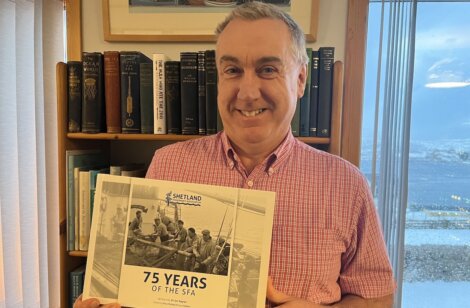 Man holding a "75 Years of the SFA" book, standing in front of a bookshelf and window.