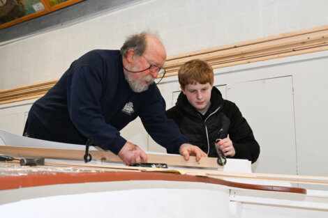 An older man instructs a younger man on woodworking, guiding him on a large piece of wood in a workshop setting.