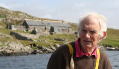 An older man stands by a lake with rocky terrain and stone houses in the background under a cloudy sky.