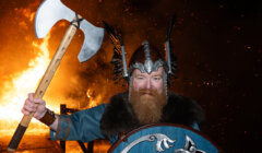 Person in Viking costume holding an axe and shield, standing in front of a large fire at night.