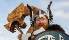 Person in Viking warrior attire with helmet and axe, positioned in front of a large wooden dragon sculpture, under a blue sky.