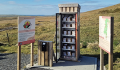 An outdoor fridge filled with pie boxes is located in a rural area, surrounded by informational signs and greenery under a blue sky.