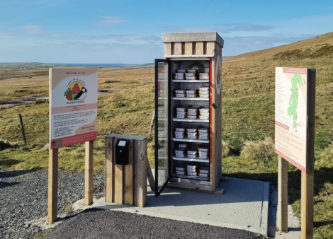 An outdoor fridge filled with pie boxes is located in a rural area, surrounded by informational signs and greenery under a blue sky.