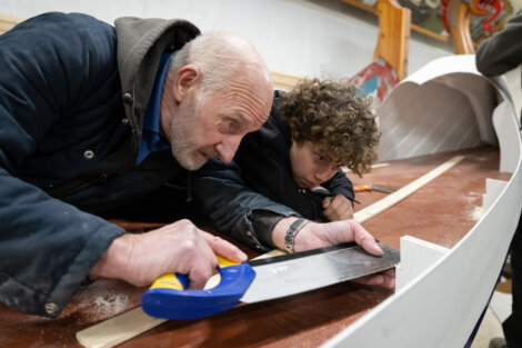 An elderly man and a young boy focus on sawing a piece of wood together on a large wooden structure indoors.