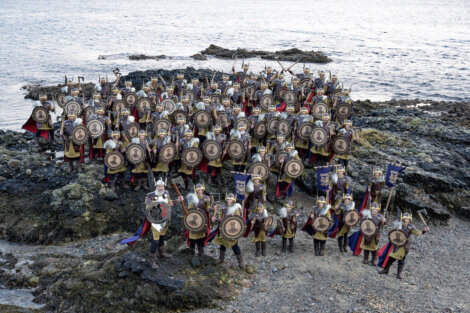 A large group of people dressed as medieval warriors stand on a rocky shoreline, holding shields and weapons.