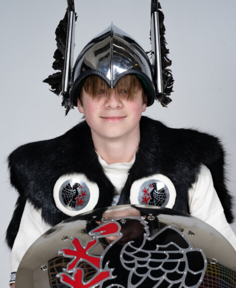 Person wearing a shiny helmet with black feathers, black fur vest, and holding a shield with a bird design.