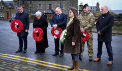 Six people stand solemnly holding wreaths of red poppies, participating in a remembrance ceremony on a wet street with stone buildings in the background.